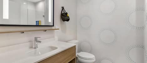 Un baño moderno con un lavamanos blanco, tocador de madera y un inodoro. Un espejo refleja la habitación, y un bolso cuelga en la pared.