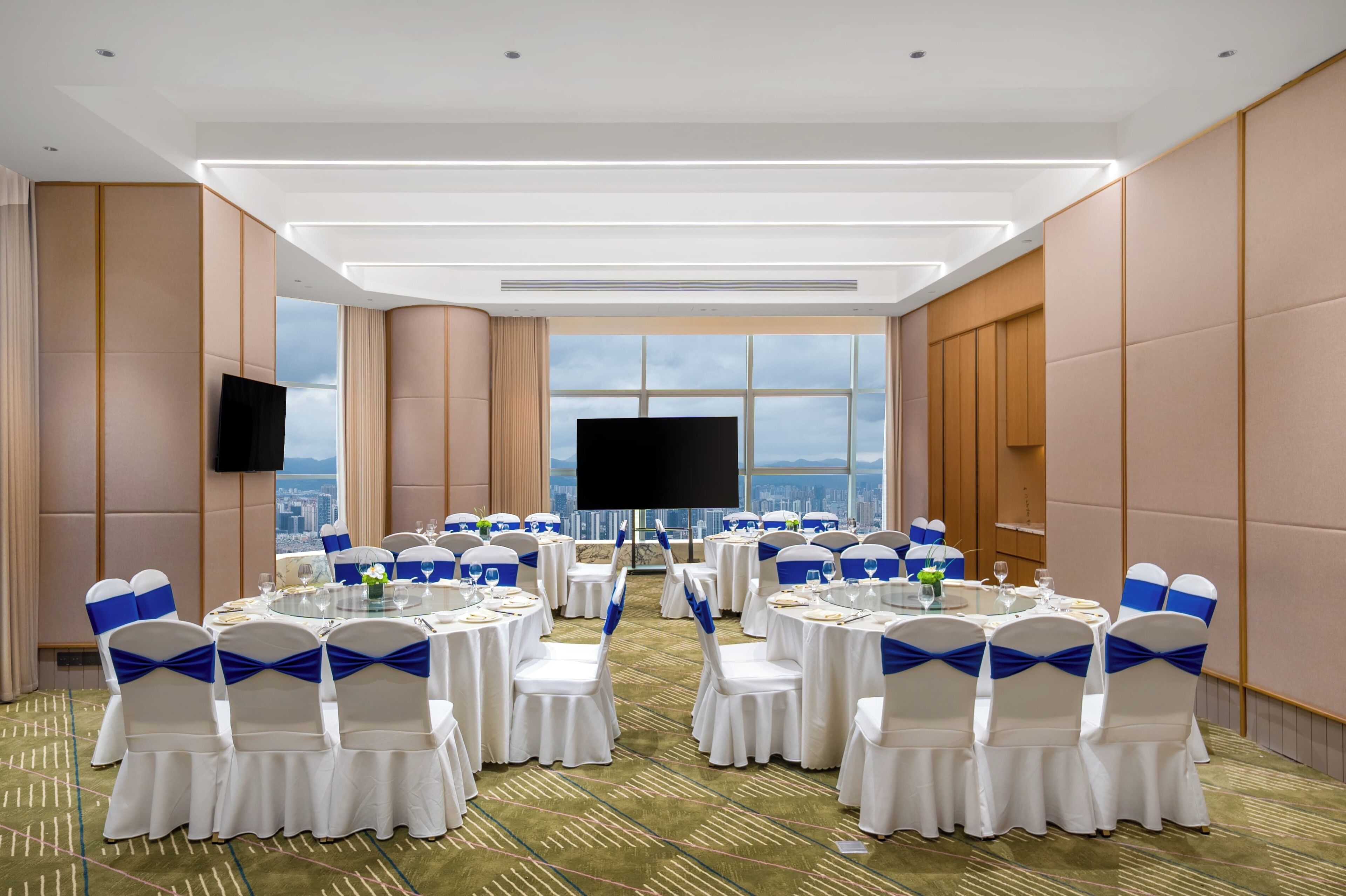 A conference room with round tables, white chairs with blue sashes, a large TV screen, and a city view through large windows.