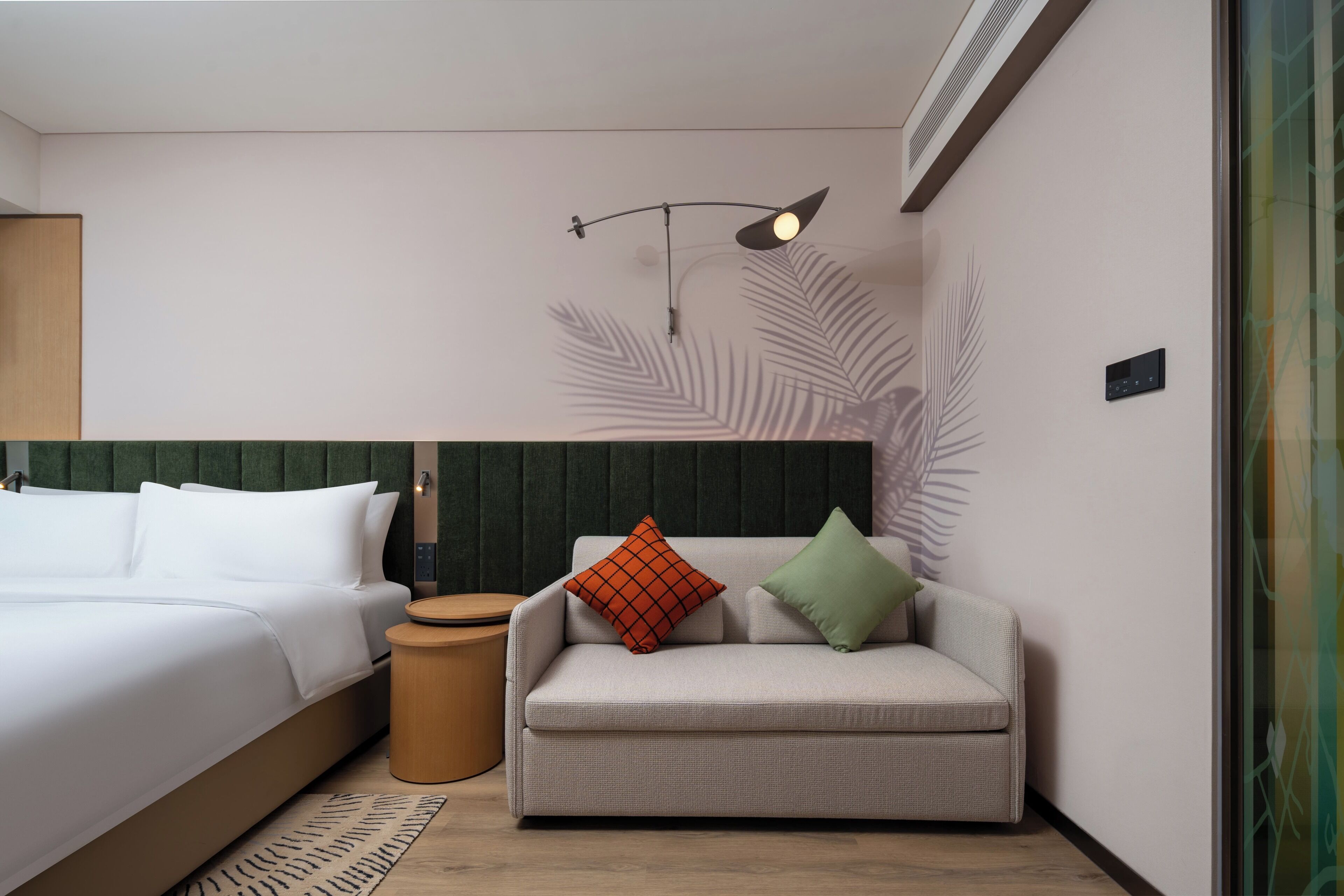 A modern hotel room with a bed, a sofa, a small table, and a wall-mounted lamp.