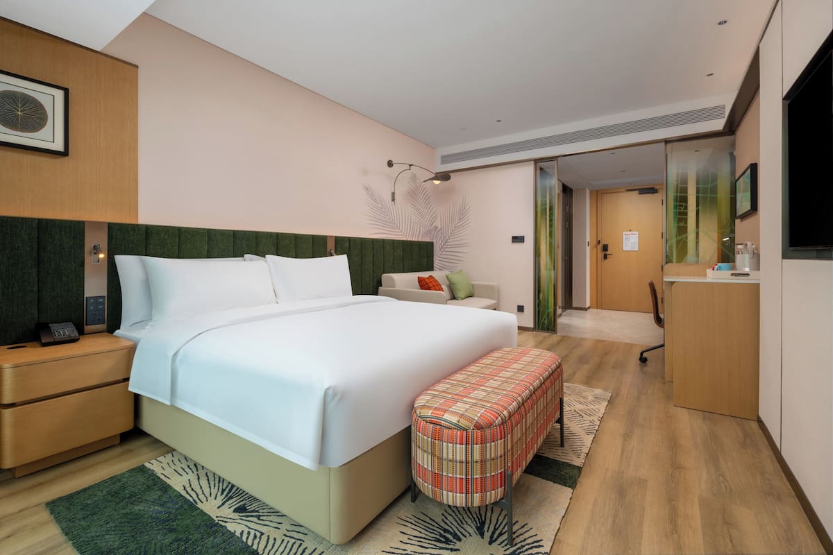 A modern hotel room with a large bed, a desk, a chair, a bench, a TV, and a door leading to another room.