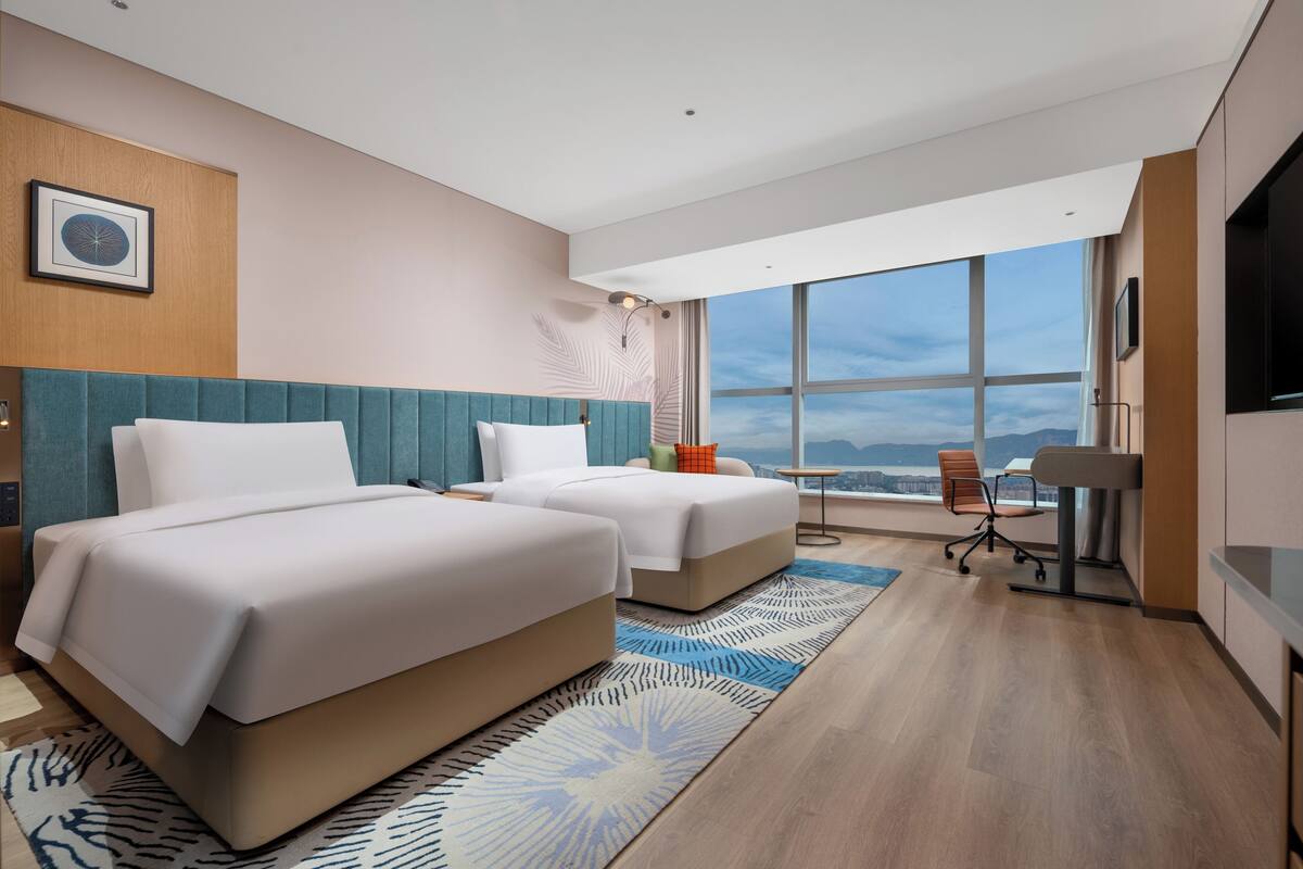 A modern hotel room with two beds, a desk, and a large window offering a city view.