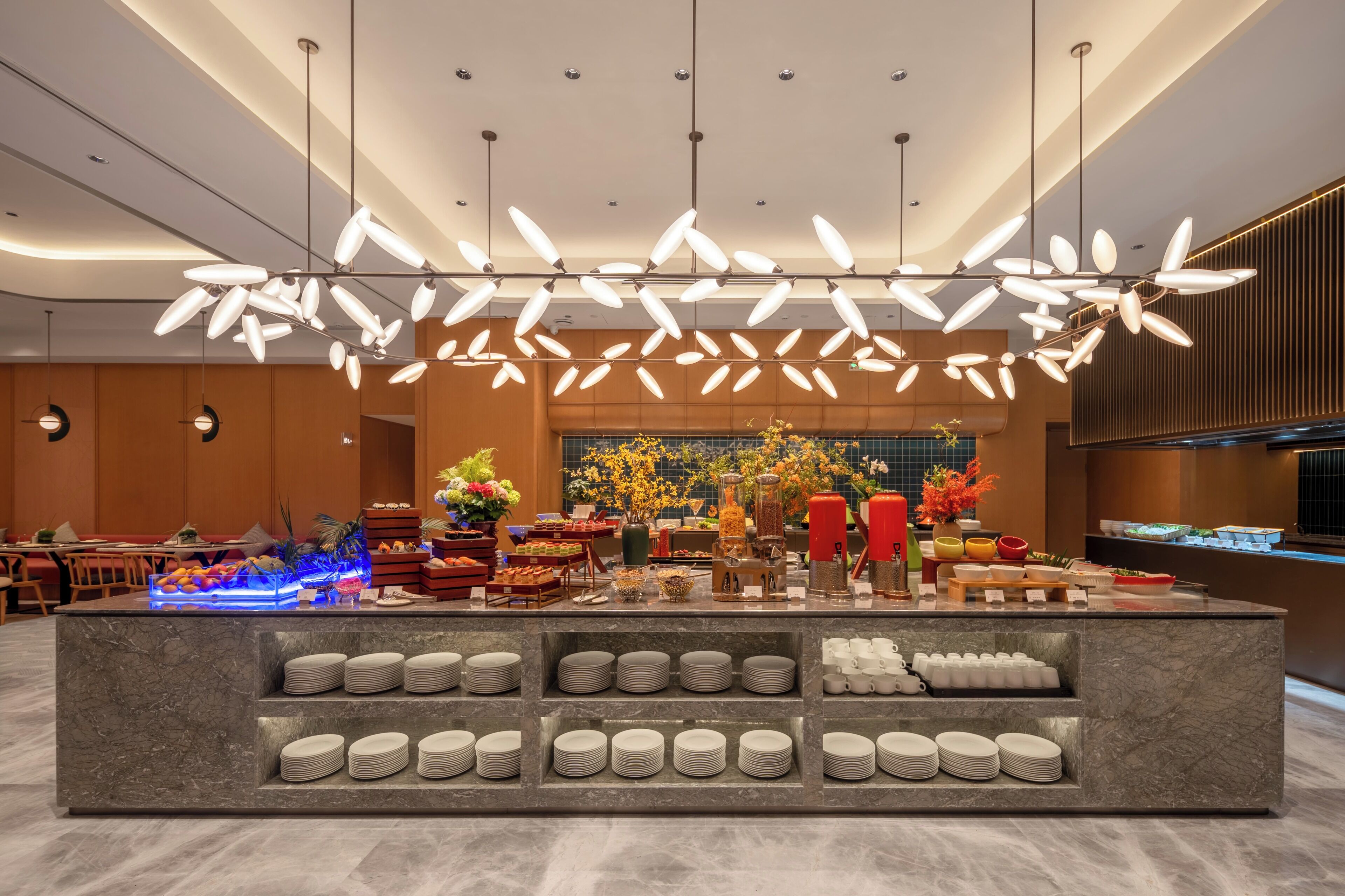 A modern hotel lobby with a marble reception desk, a variety of food and drink stations, and a large, decorative light fixture above.