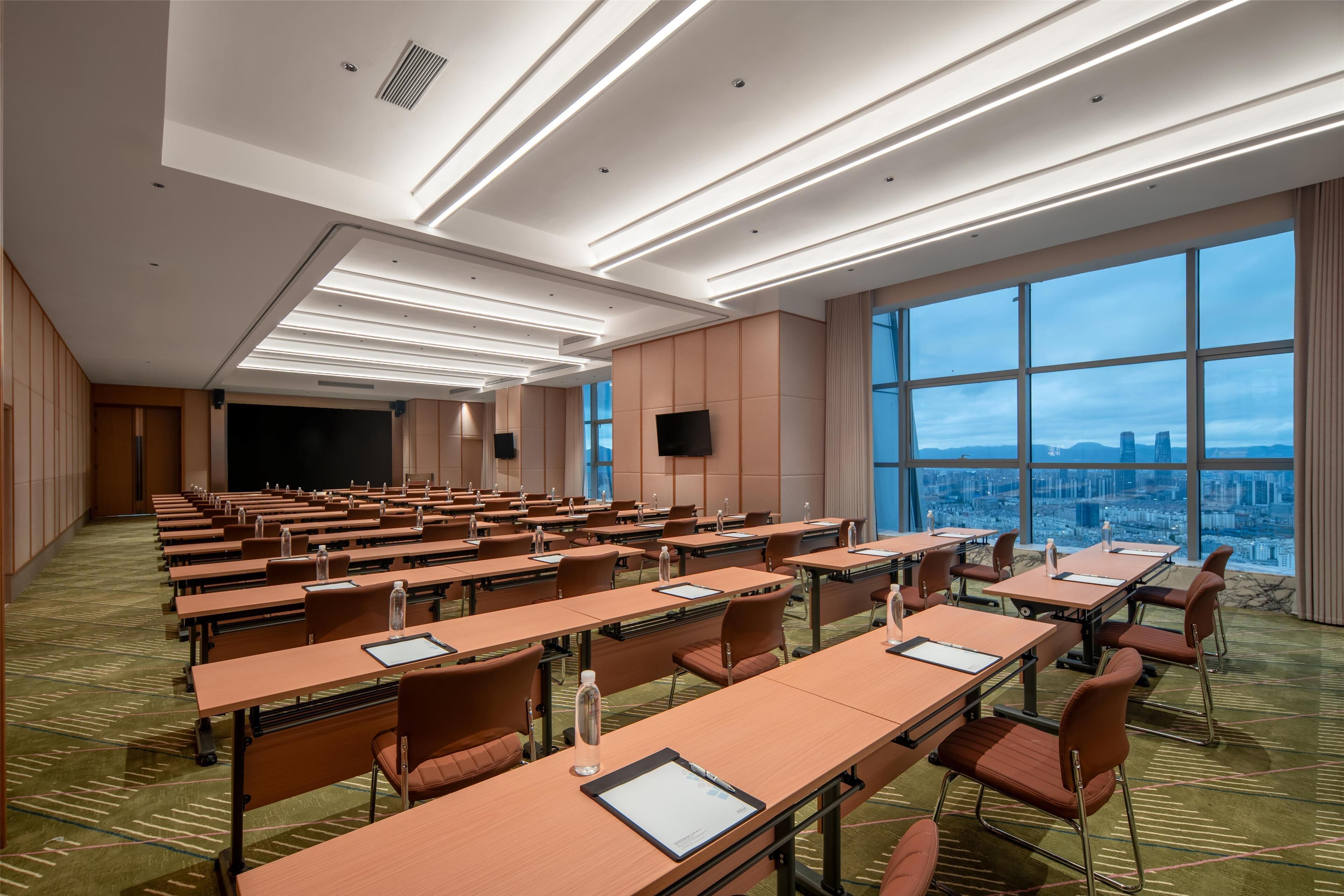 A conference room with rows of tables and chairs, large windows with a city view, and modern lighting fixtures.