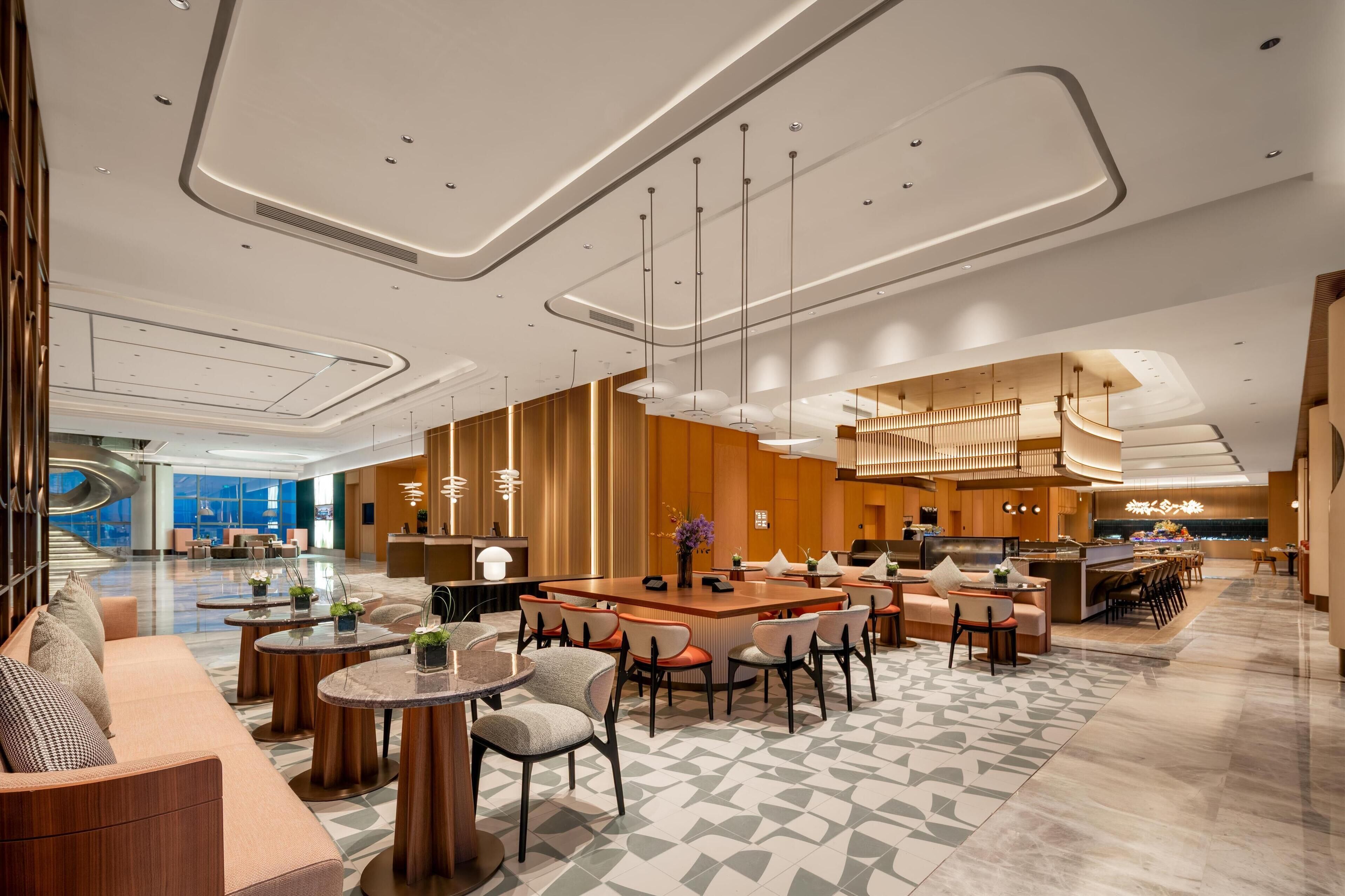 A modern hotel lobby with a central seating area, a reception desk, and a kitchen visible in the background.