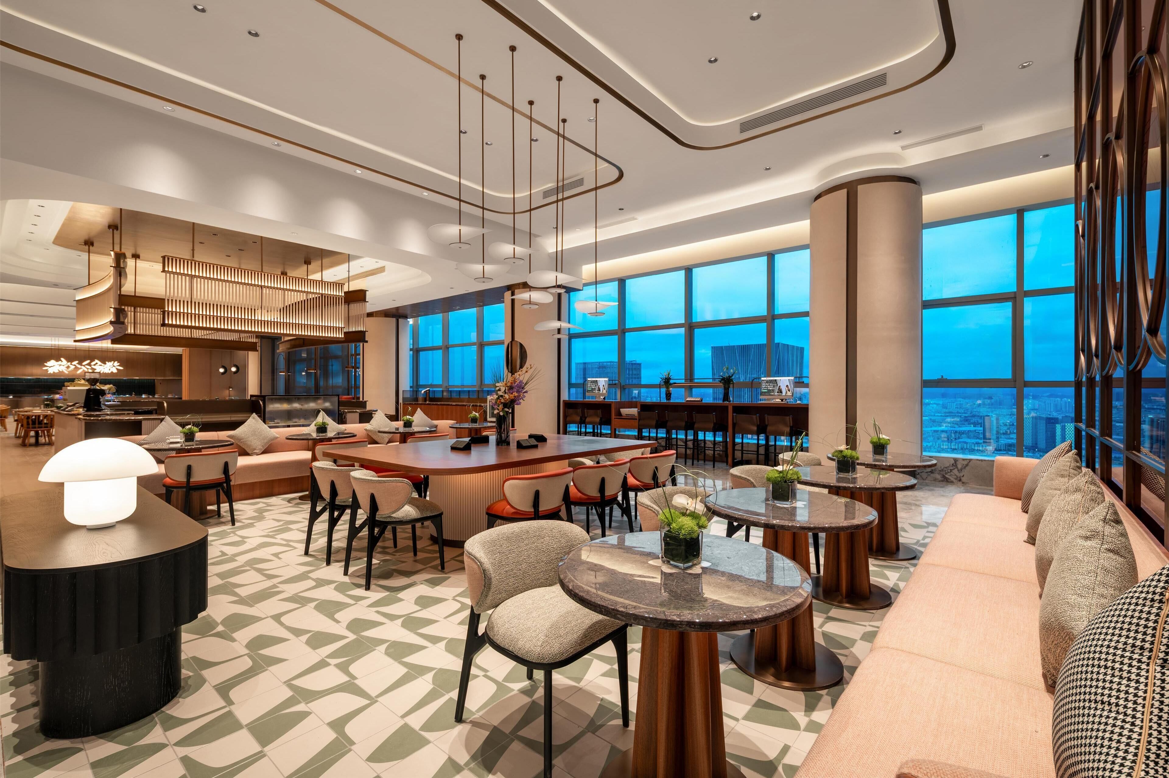 A modern hotel lobby with a bar, seating areas, and large windows offering city views.