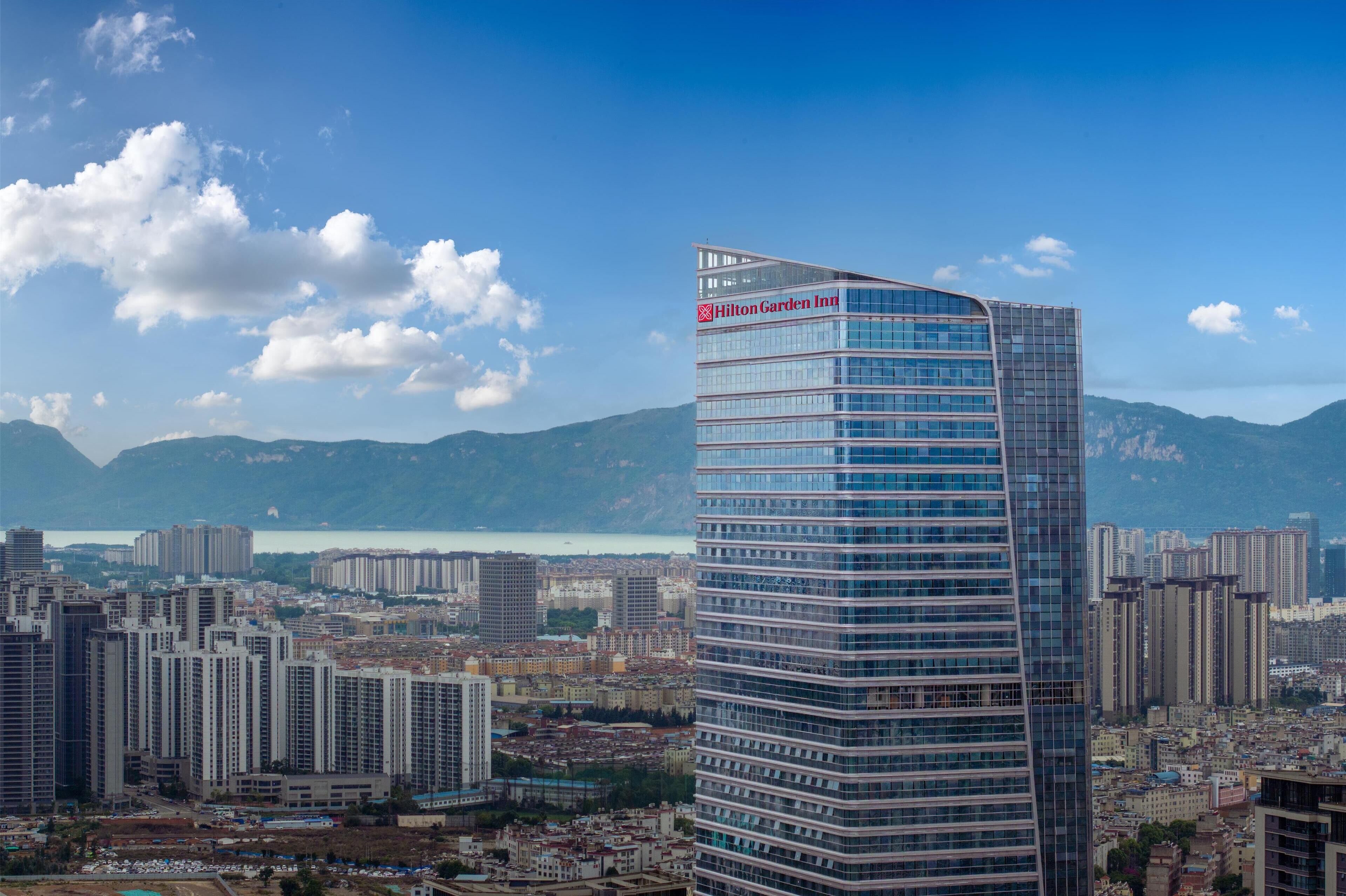 Un hotel de gran altura con fachada de vidrio moderna y el logotipo visible de Hilton Garden Inn, rodeado por un paisaje urbano y montañas al fondo.