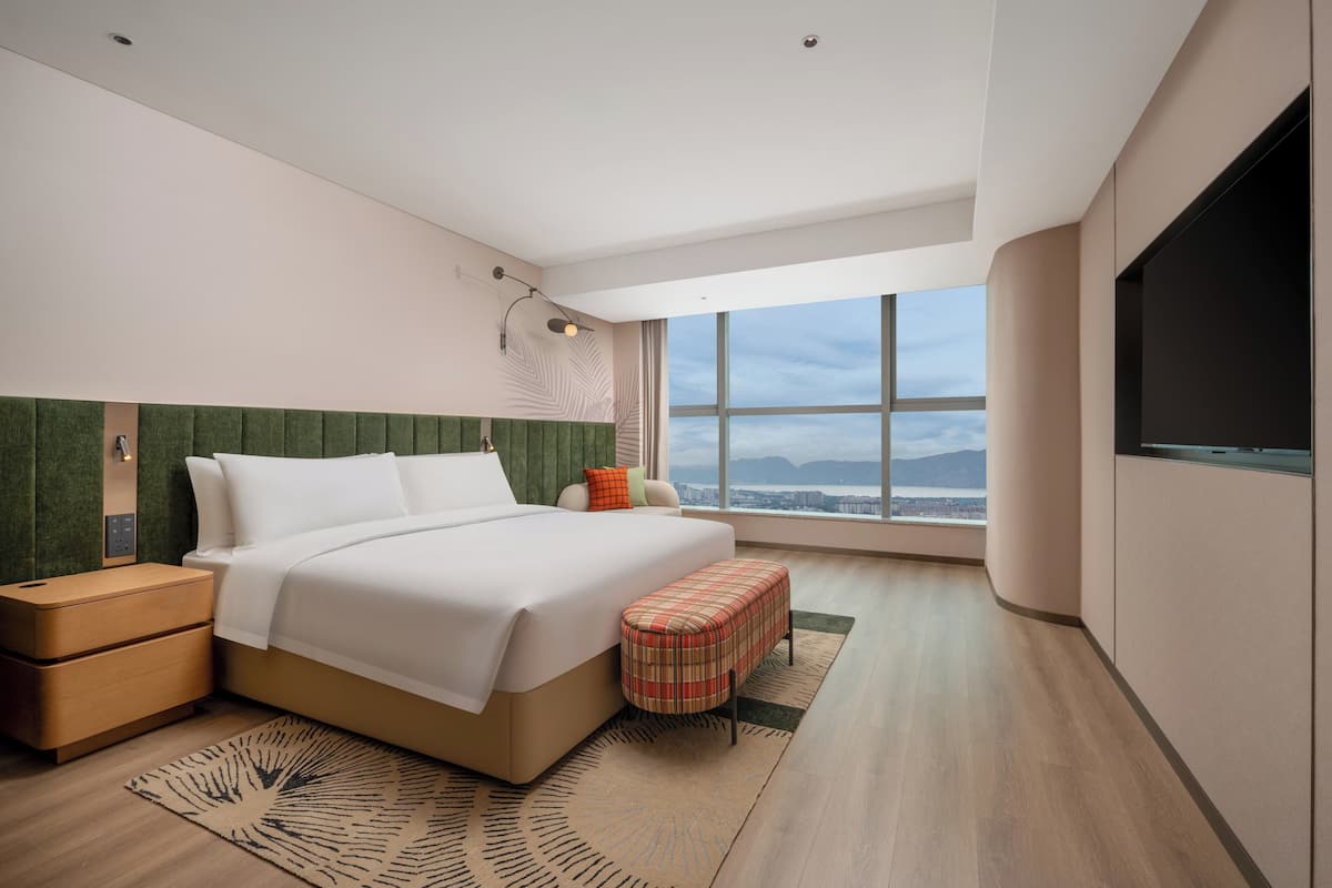 A modern hotel room with a large bed, a nightstand, a bench, a TV, and a large window offering a city view.