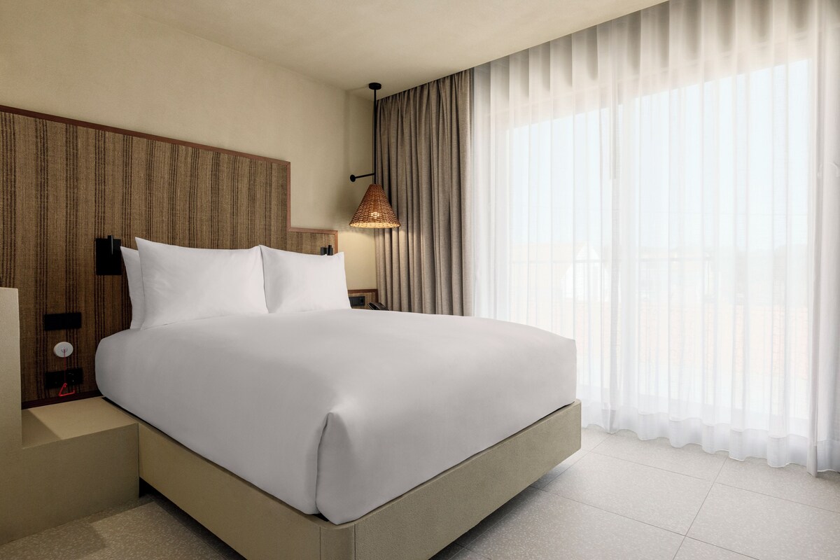 Une chambre d’hôtel moderne avec un grand lit, une tête de lit en bois, une table de chevet avec une lampe et des rideaux légers à la fenêtre.