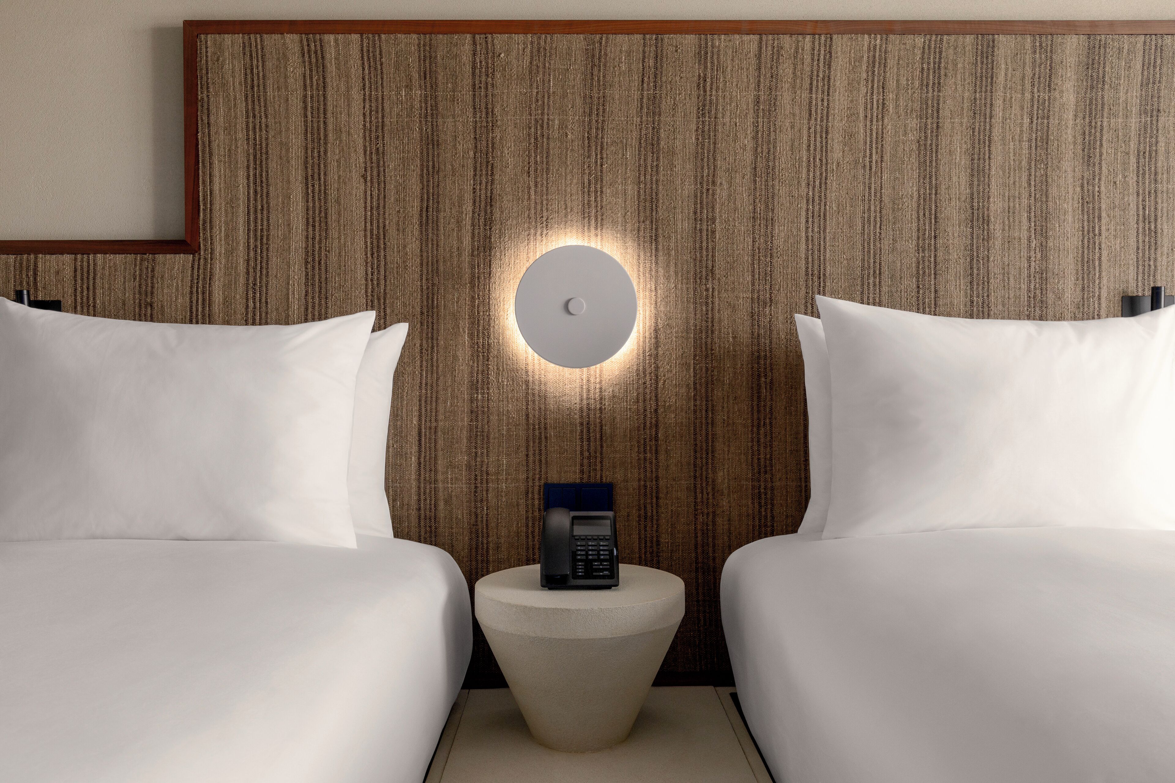 Quarto de hotel com duas camas, uma mesa redonda com um telefone e um candeeiro fixado na parede.