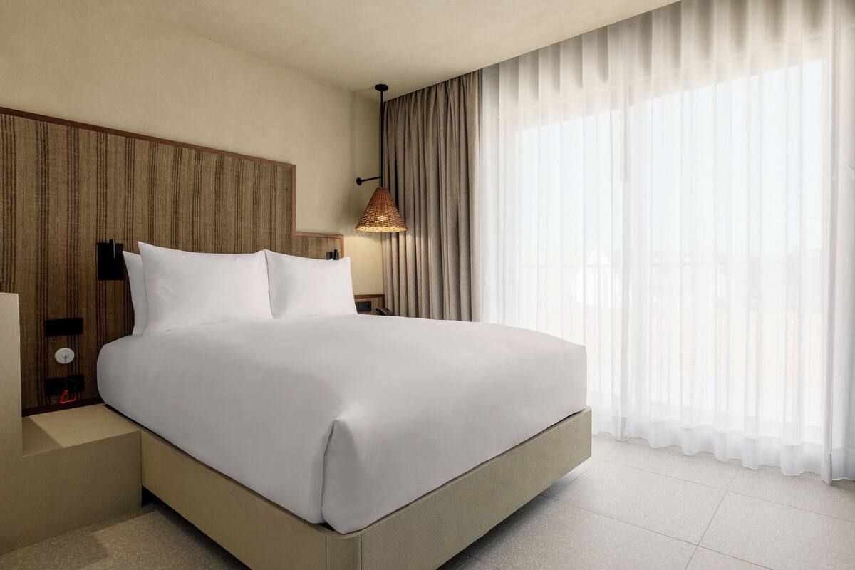 Une chambre d’hôtel moderne avec un grand lit, une tête de lit en bois, une table de chevet avec une lampe et des rideaux légers à la fenêtre.
