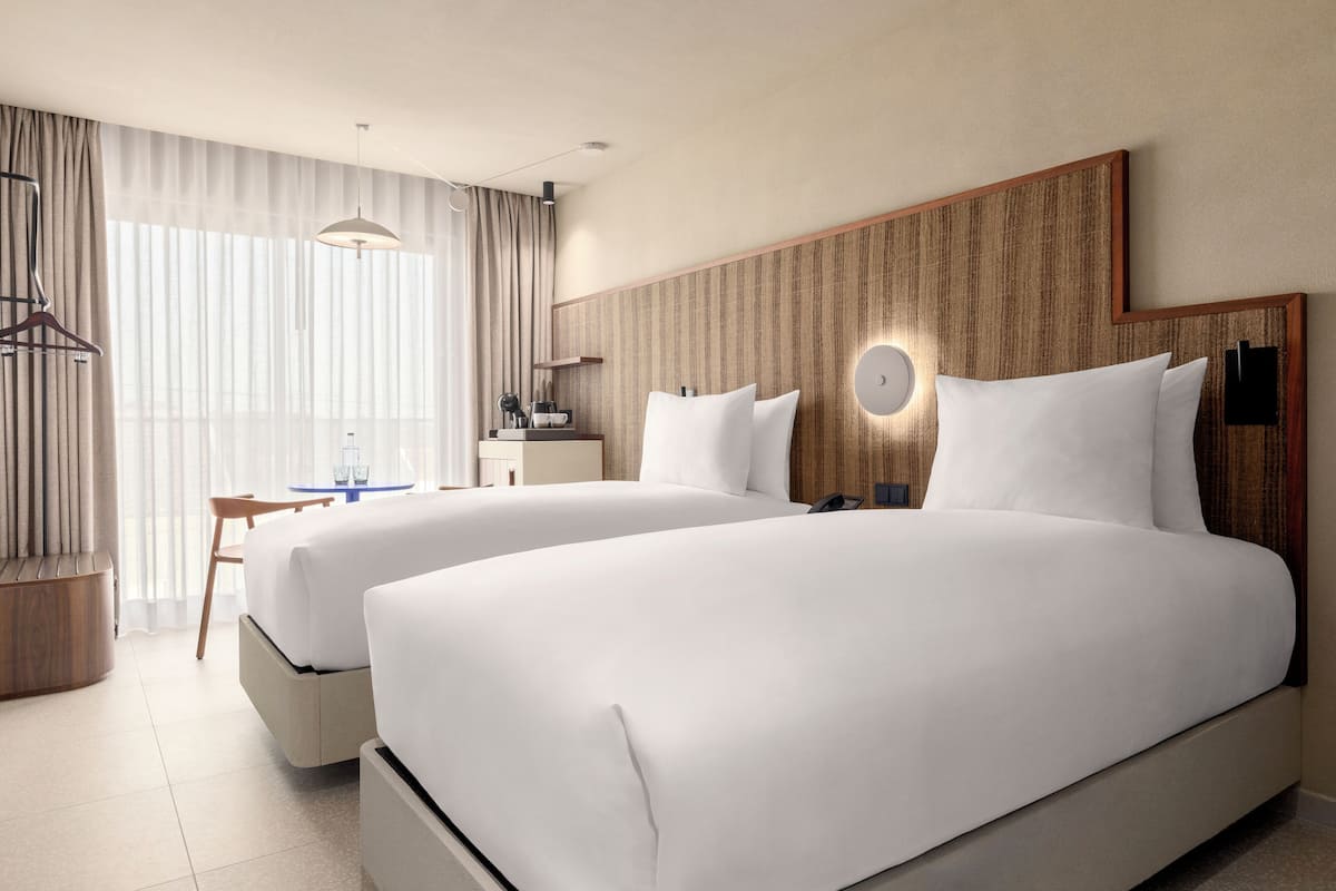 Une chambre d’hôtel avec deux lits, un bureau, une chaise, un luminaire suspendu et une grande fenêtre avec des rideaux.