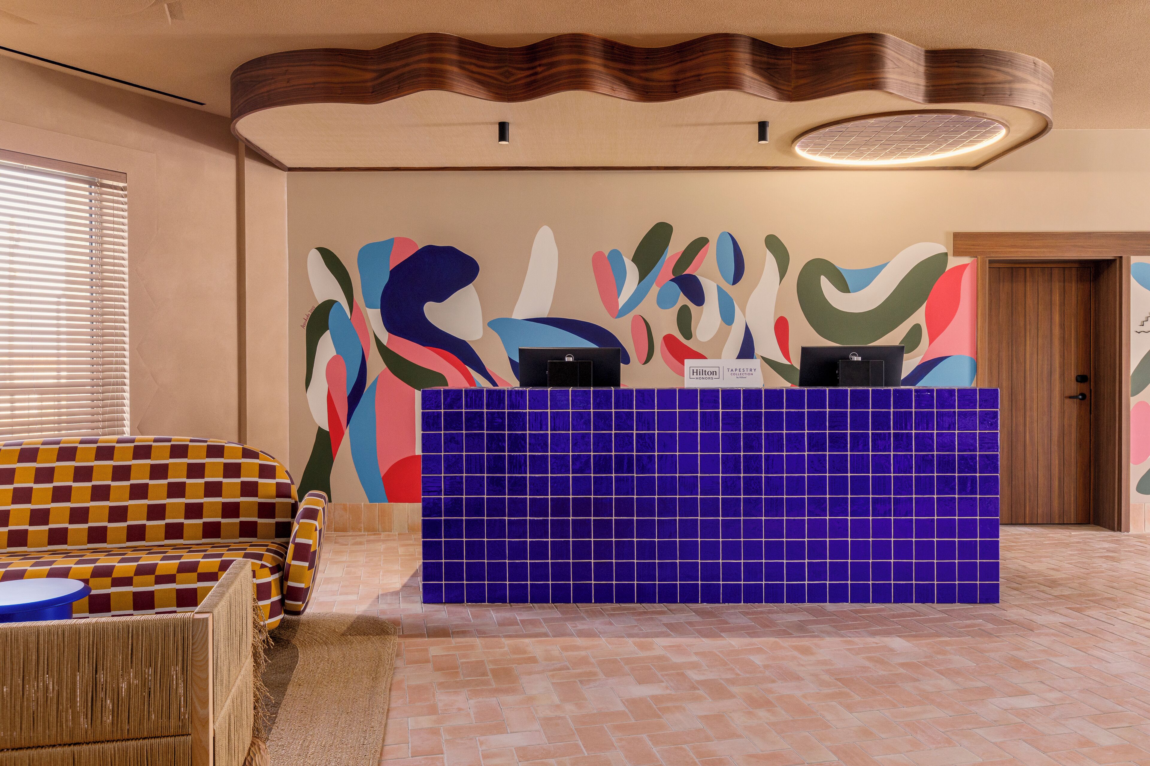 Zona de recepció amb un taulell d'enregistrament, una paret amb un disseny abstracte i colorit, un sofà amb dibuixos i una porta de fusta.
