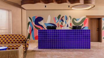 A reception area with a check-in counter, a colorful abstract wall, a patterned sofa, and a wooden door.