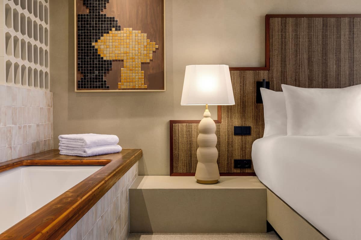Une chambre d’hôtel avec une baignoire en bois, un lit blanc, une lampe fixée au mur et une œuvre d’art en mosaïque.