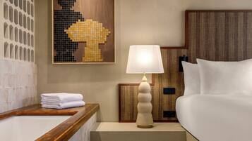 A hotel room with a wooden bathtub, a white bed, a wall-mounted lamp, and a mosaic artwork.