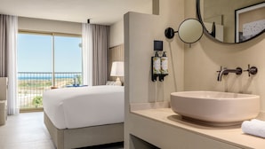 Habitación de hotel con una cama grande, un baño con un espejo redondo y vista al océano por la ventana.