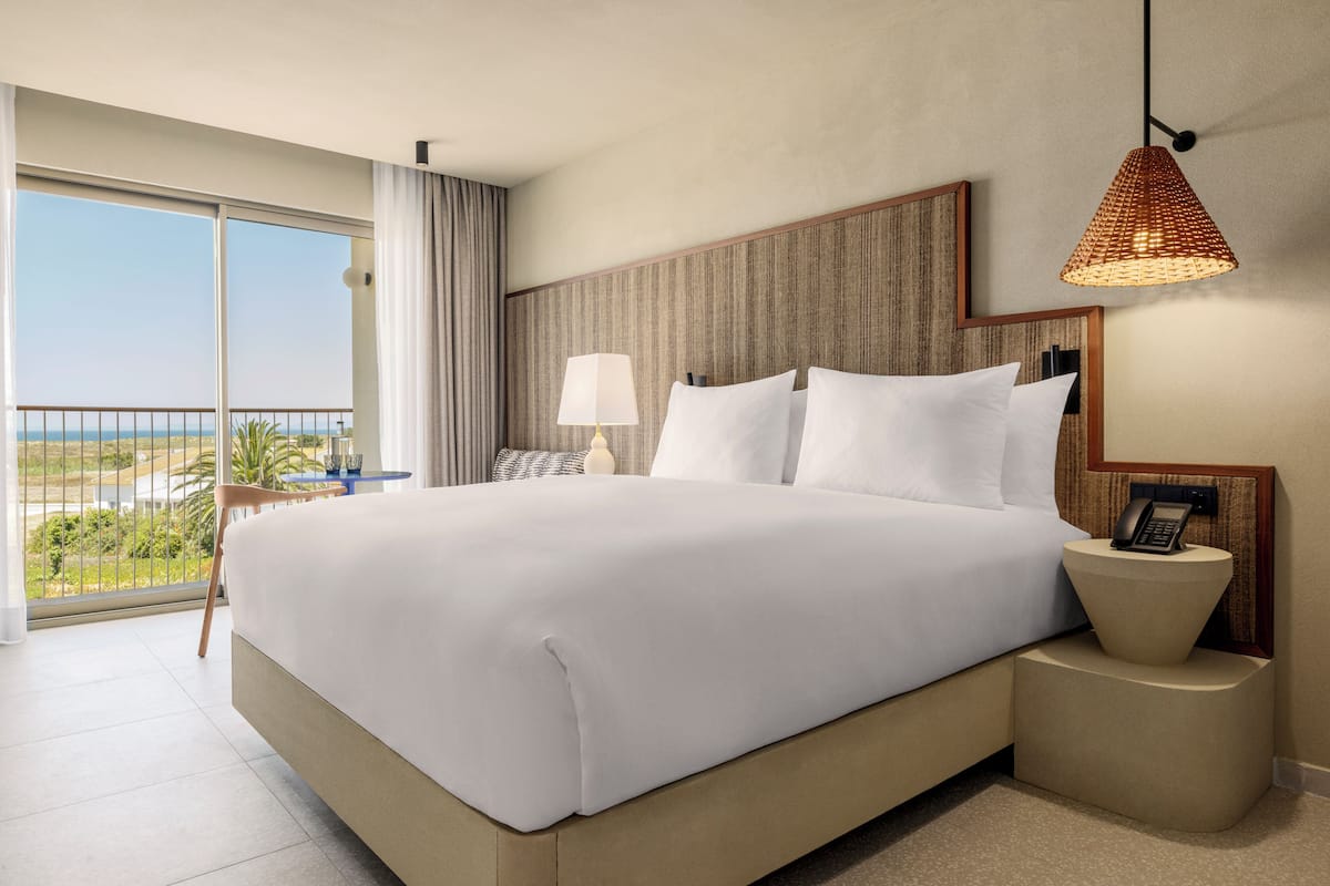 Une chambre d’hôtel avec un grand lit, une table de chevet avec une lampe, un téléphone et une vue sur l’océan depuis un balcon.