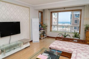 A living room with a TV, a glass coffee table, a bed with floral bedding, a white cabinet, and a large window with a view of the sea. - Jinshatan Family Hostel (Qingdao)