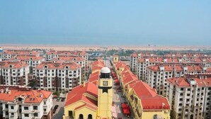 Un edificio alto con una torre central, rodeado de otros edificios y una playa al fondo.