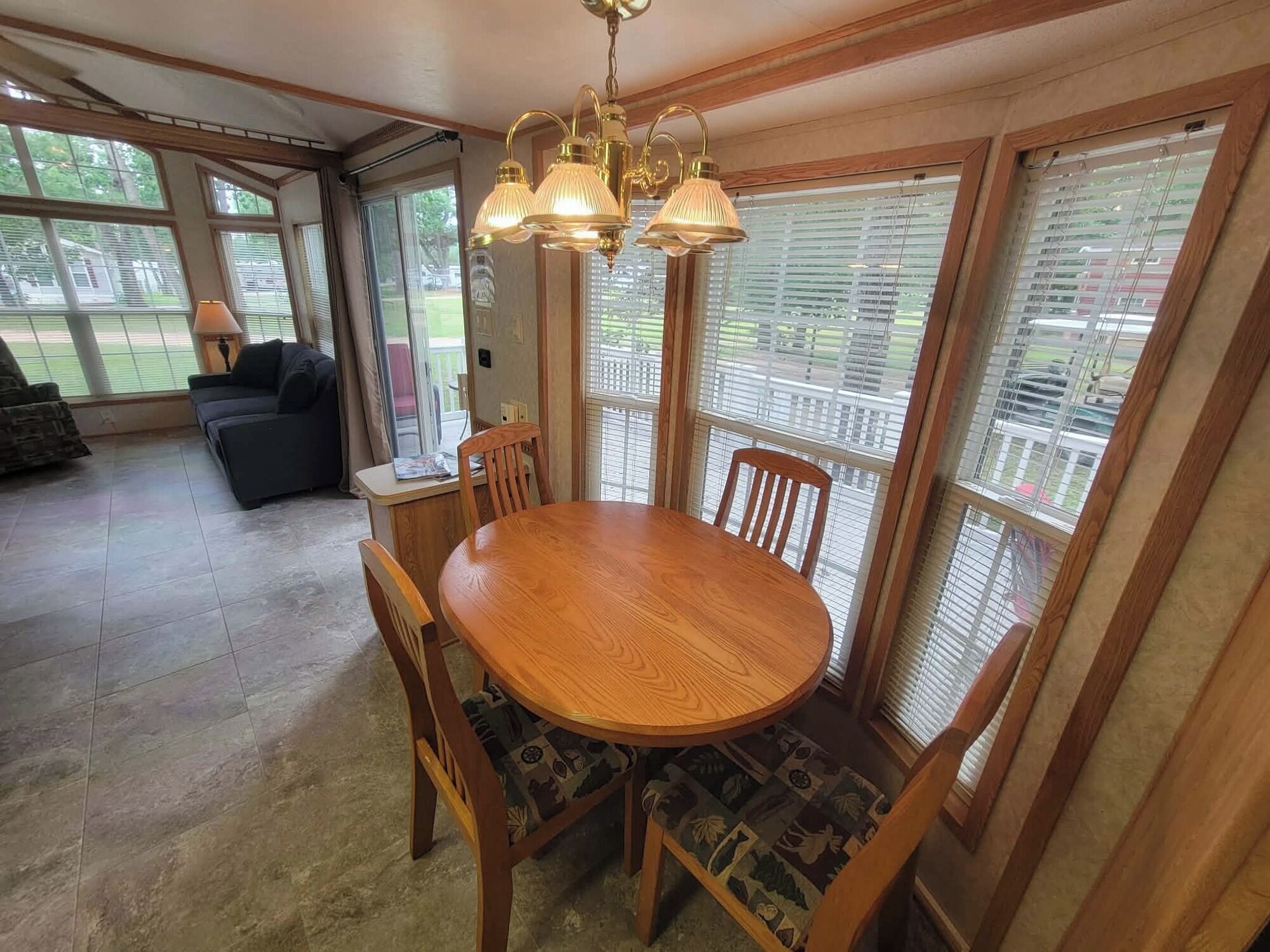 A dining area with a wooden table and chairs, a chandelier, and large windows with blinds.