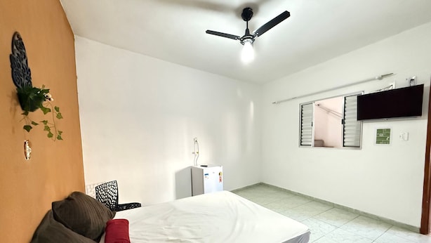 Une chambre avec un lit, un ventilateur de plafond, un téléviseur fixé au mur et un petit réfrigérateur.