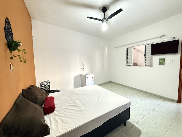 A bedroom with a bed, a ceiling fan, a TV mounted on the wall, and a small refrigerator. - Pé na Areia Praia da Aviação II (Praia Grande)