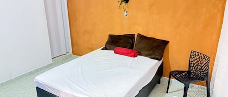 A bed with a white mattress, two dark pillows, and a red accent pillow. A black chair is positioned to the right of the bed.