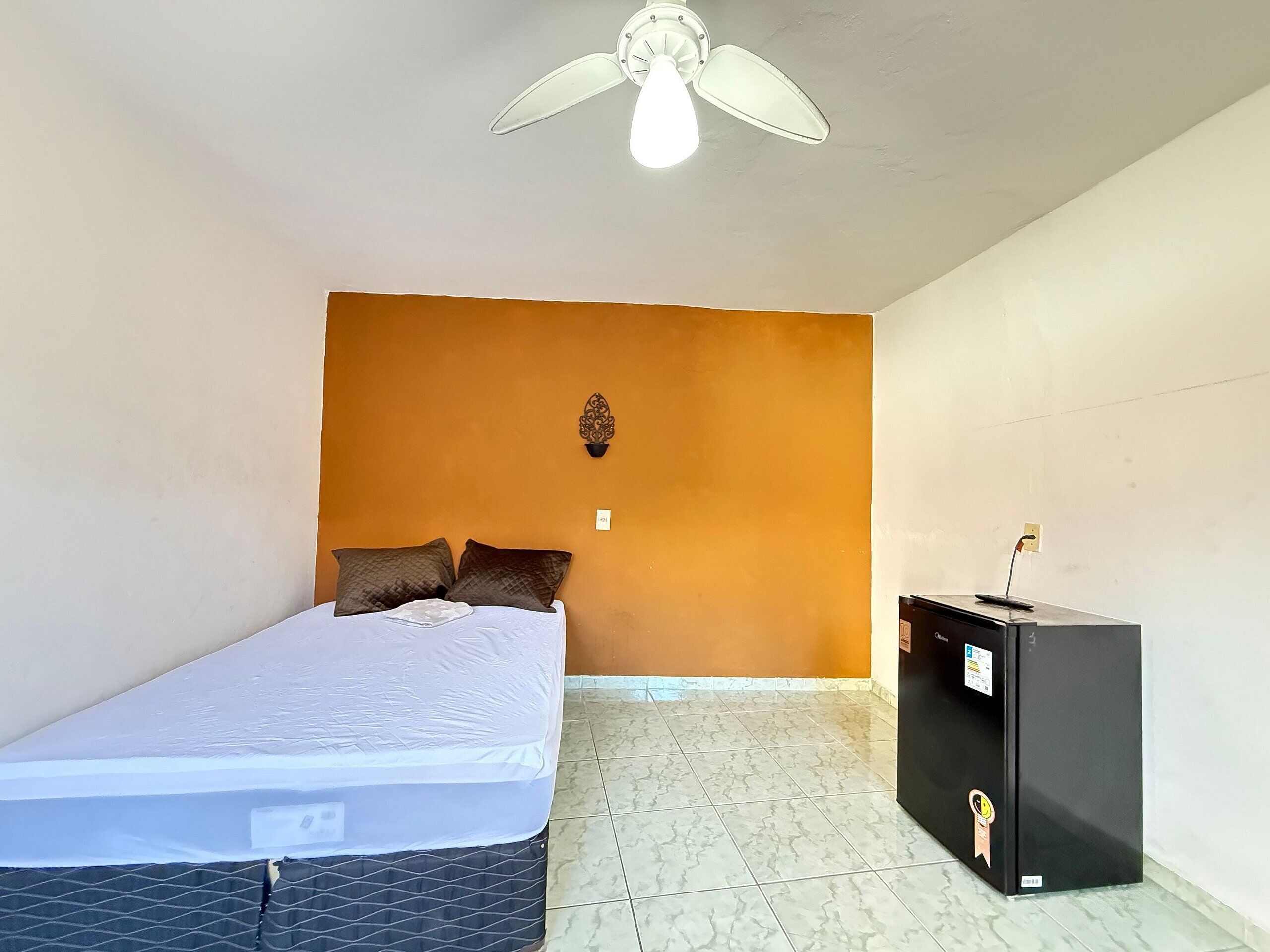 Una habitación pequeña con cama individual, pared de acento naranja, ventilador de techo y minibar negro.