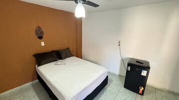 A bedroom with a bed, a ceiling fan, a wall-mounted light, and a black box on the floor.
