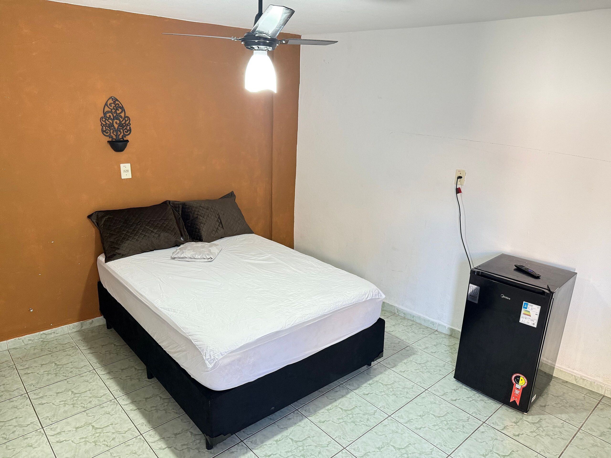 Ein Schlafzimmer mit einem Bett, einem Deckenventilator, einem kleinen schwarzen Schrank und einer Wandsteckdose.