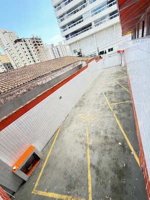 A parking area with marked spaces, a fenced walkway, and a building with multiple floors in the background. - Pé na Areia Praia da Aviação II (Praia Grande)