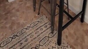 Una cocina con una mesa de madera oscura y dos taburetes, un cuadro en la pared y una ventana con cortinas.