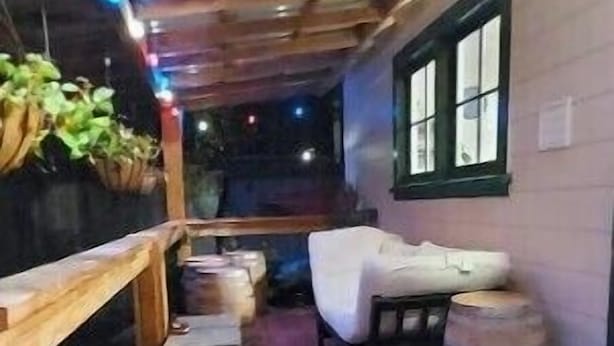 Una terraza de madera con un sofá y un barril, rodeada de muros y una ventana.