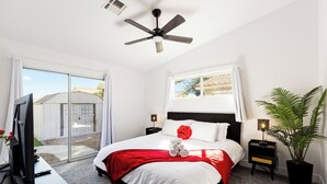 A bedroom with a bed, a TV, a ceiling fan, a window, a nightstand, and a potted plant. - Modern Oasis: Cozy Retreat Near the Strip (North Las Vegas)