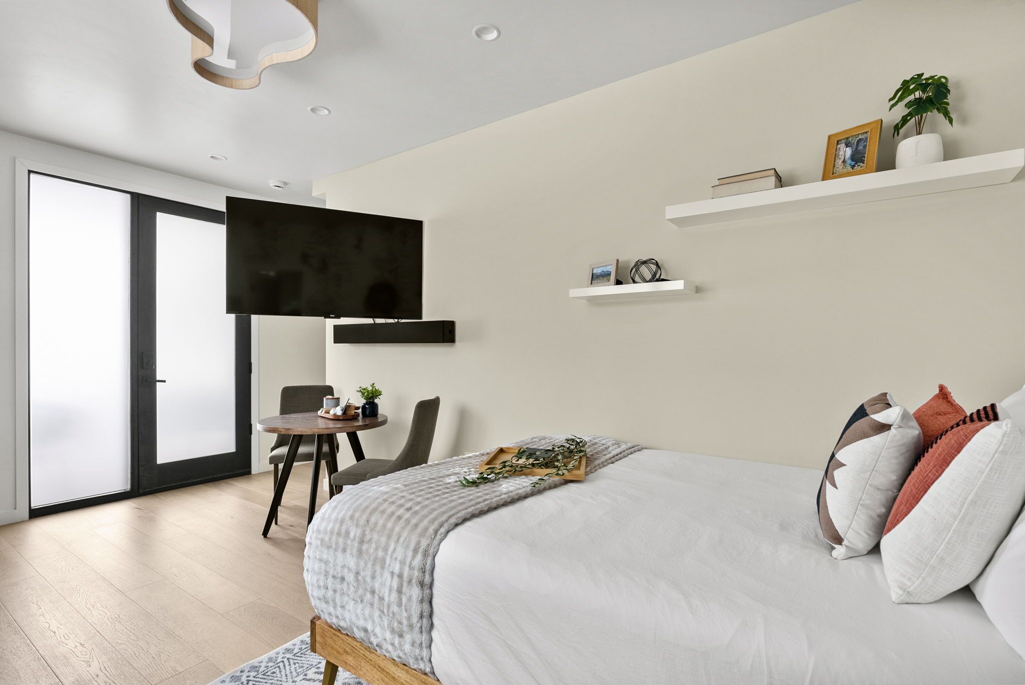 A modern bedroom with a large bed, a flat-screen TV mounted on the wall, a small table with a chair, and a sliding glass door leading to a balcony.