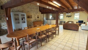 Uma sala de jantar espaçosa com uma mesa de madeira comprida e cadeiras, uma cozinha ao fundo e uma sala de estar com sofá e televisão.
