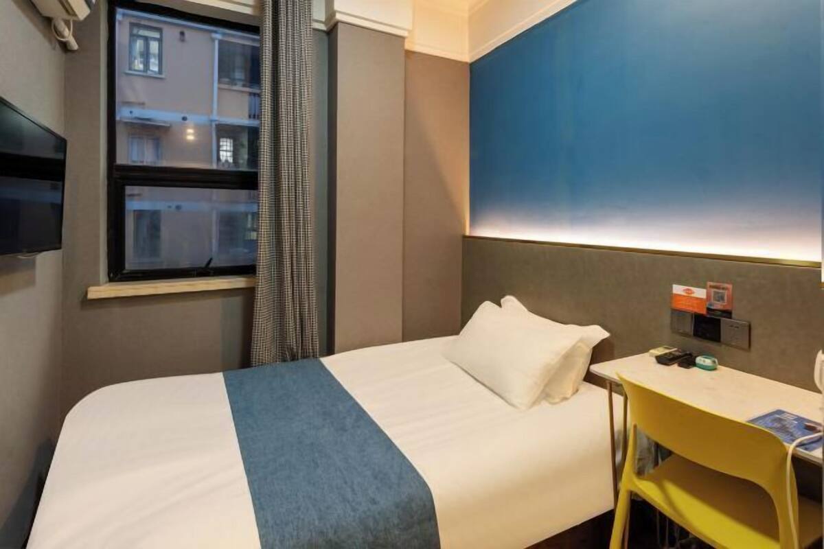 A hotel room with a bed, a desk, a chair, a window with curtains, and a wall-mounted TV.