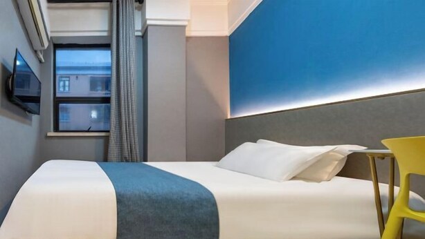 Habitación de hotel con cama, televisor en la pared, ventana con cortinas y vistas al exterior.