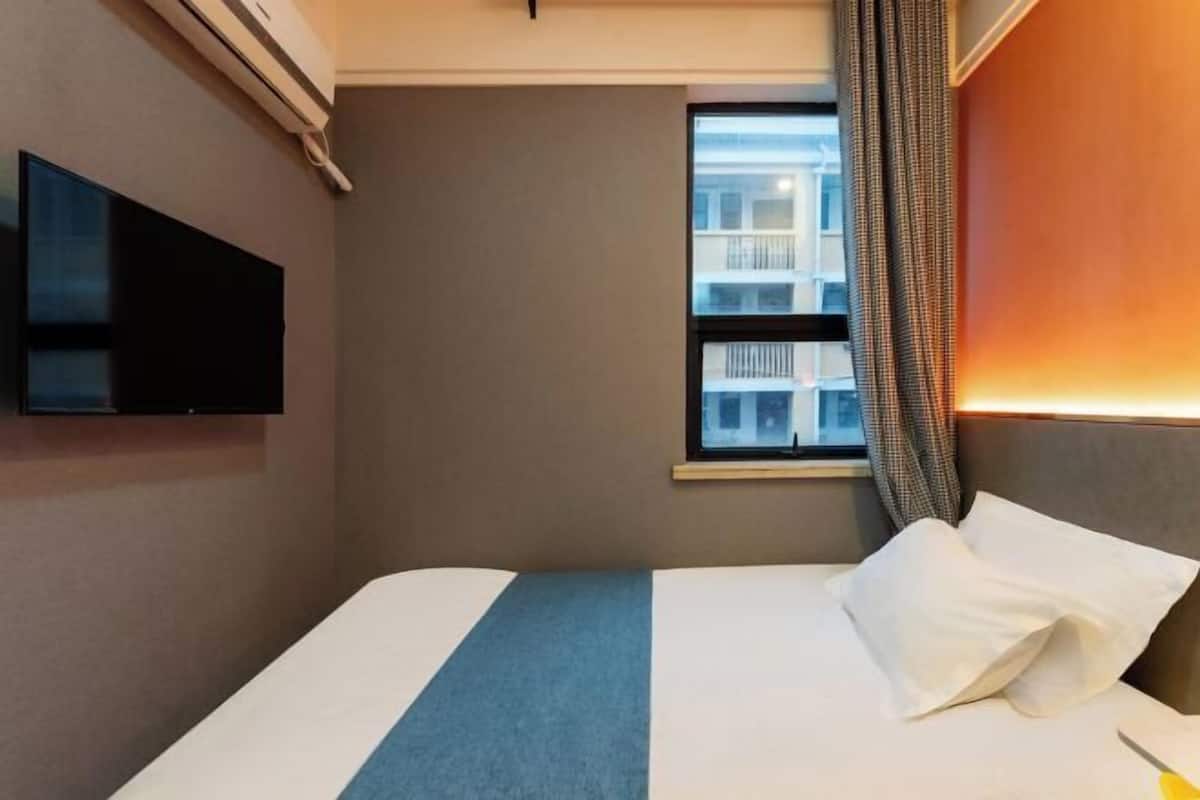 A hotel room with a bed, a flat-screen TV mounted on the wall, a window with curtains, and a view of buildings outside.
