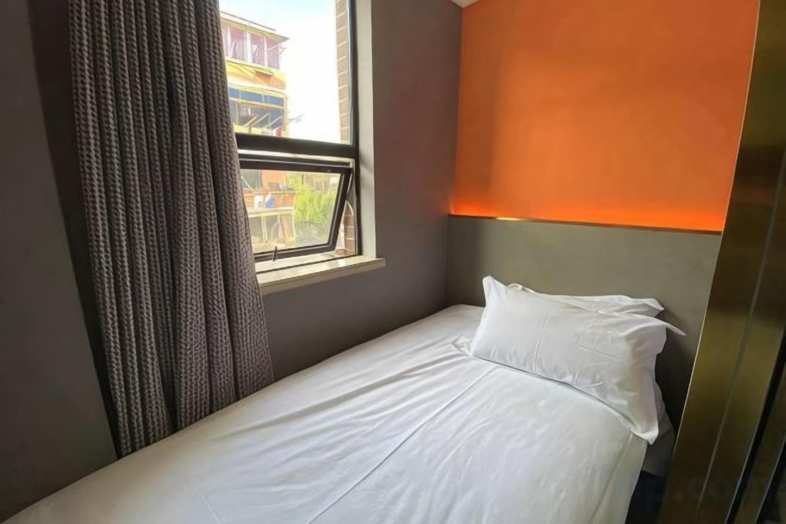 A single bed with white bedding in a room with a window, a curtain, and a wall-mounted light fixture.