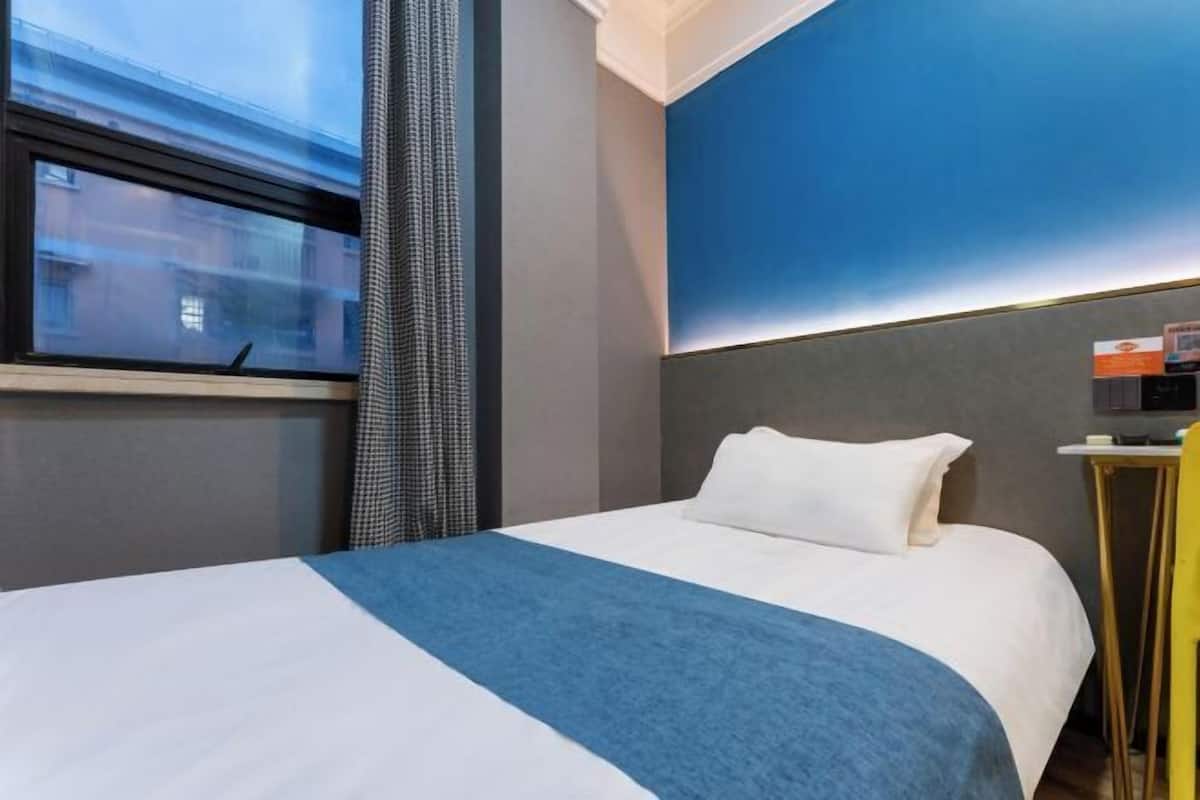 A hotel room with a bed, a window, a curtain, a bedside table, and a wall with a blue accent.