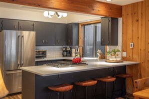 A modern kitchen with dark cabinets, stainless steel appliances, a central island with stools, and wooden accents. - Sunriver Nostalgia - Walk to Sunriver Village, SHARC passes, HOT TUB (Sunriver)