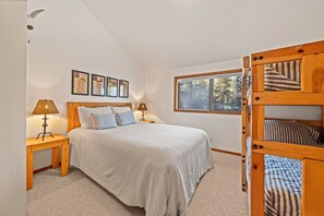 A bedroom with a large bed, two bedside tables with lamps, a window with a view of trees, and a bunk bed structure.