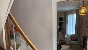 Un escalier avec des rampes en bois et des balustrades blanches mène à un salon agrémenté d’un tapis à motifs, d’un fauteuil vert et d’une fenêtre avec des rideaux.