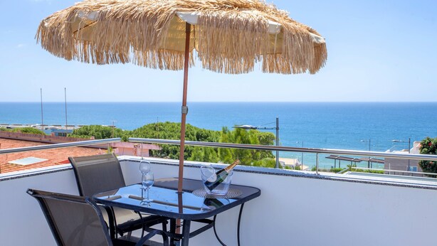 Un balcon avec une table, des chaises, un parasol en paille et une vue sur la mer.