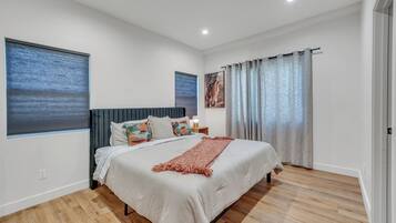 A modern bedroom with a large bed, wooden flooring, a headboard, a nightstand, and a window with blinds.