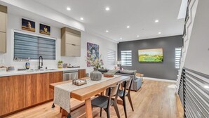 Een moderne keuken met houten kastjes, een zithoek met een houten tafel en stoelen, en een groot raam met jaloezieën.