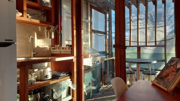 Een keuken met houten planken, een gootsteen en een fornuis. Er is een raam met uitzicht op bomen en een tafel met een stoel.