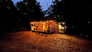 Een klein, verlicht huisje met een veranda en een deur, omringd door bomen.