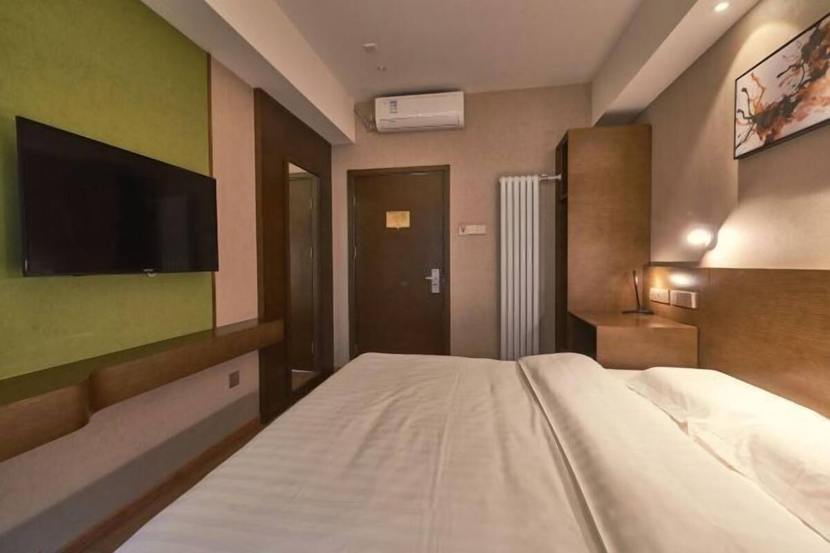 A hotel room with a bed, a flat-screen TV mounted on the wall, a bedside table with a lamp, and a door with a light switch.